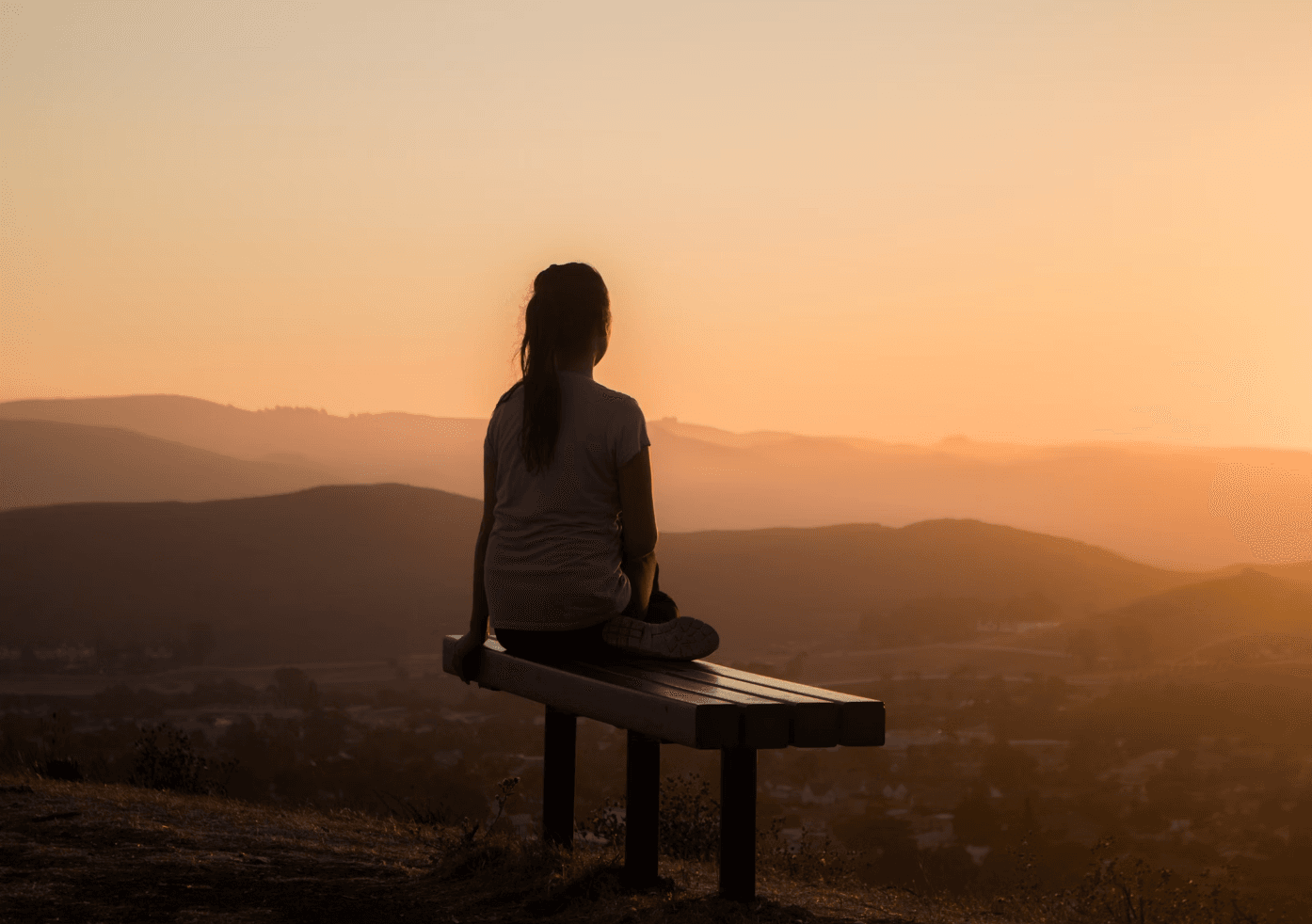 woman sitting on a bench and looking at the sunset over the horizon of the mountain tops