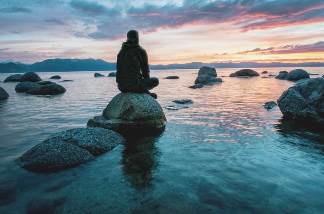 person sitting on a rock on a body of water looking into a pink sunset